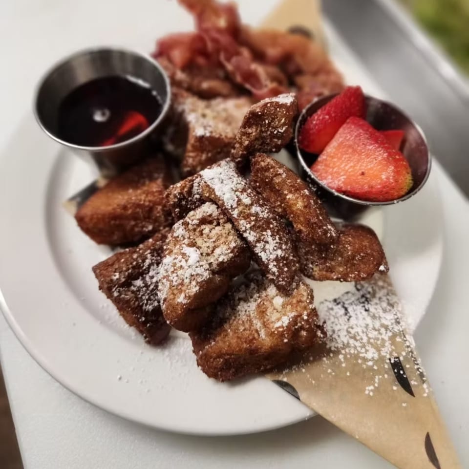 Astrid's French toast bites with powdered sugar, bacon, strawberries, and syrup on a white plate.