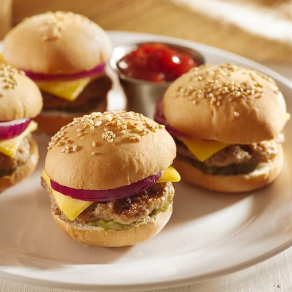 Four mini cheeseburger sliders with red onion and pickles, served with a side of ketchup.
